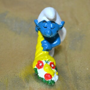 May include: A blue Smurf figurine with a white hat and a yellow cornucopia. The cornucopia is decorated with red and white flowers and green leaves. The figurine is a collectible item.
