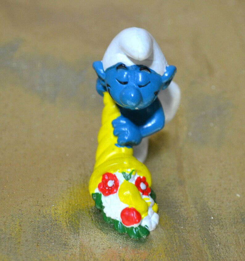 May include: A blue Smurf figurine with a white hat and a yellow cornucopia. The cornucopia is decorated with red and white flowers and green leaves. The figurine is a collectible item.