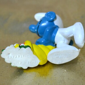 May include: A blue and white Smurf figurine with a yellow base, holding a floral arrangement. The figurine is lying on its side, with the text "&copy; 1976 PEYO SCHLEICH" visible on the base. The figurine is made in Hong Kong.