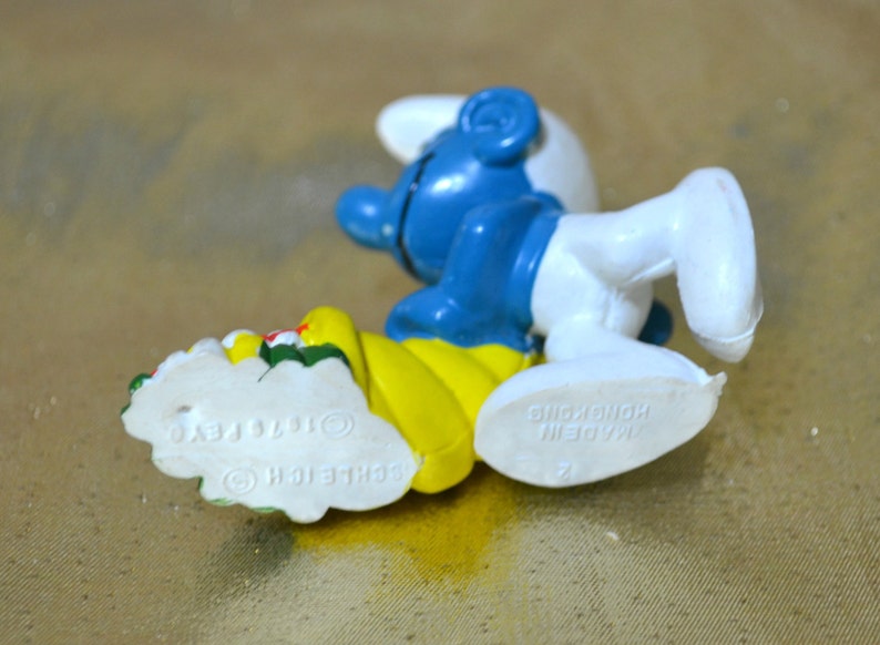 May include: A blue and white Smurf figurine with a yellow base, holding a floral arrangement. The figurine is lying on its side, with the text "&copy; 1976 PEYO SCHLEICH" visible on the base. The figurine is made in Hong Kong.