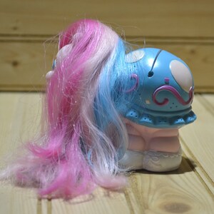 May include: A toy pony with a blue head and pink, white, and blue hair. The pony has a light pink body and white shoes. The head has white spots and pink swirls. The pony is on a wooden surface.
