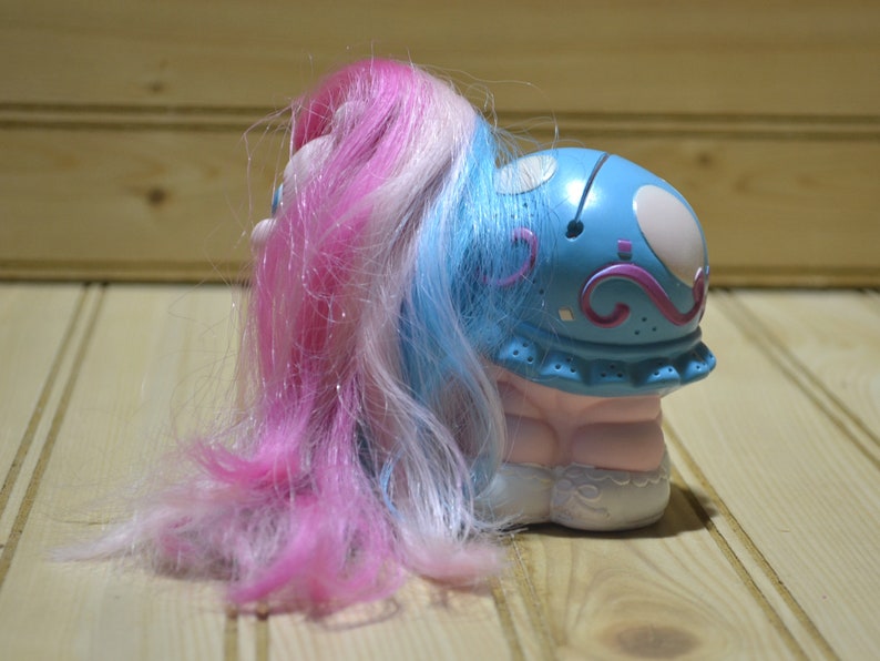 May include: A toy pony with a blue head and pink, white, and blue hair. The pony has a light pink body and white shoes. The head has white spots and pink swirls. The pony is on a wooden surface.