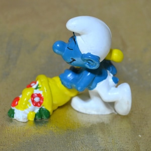 May include: A figurine of a Smurf, a blue cartoon character, running with a yellow cornucopia filled with red and white flowers. The Smurf is wearing a white hat and pants.