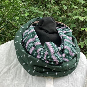 Könnte beinhalten: Ein grüner und lilafarbener, gemusterter Infinity-Schal mit weißem Blumenmuster. Der Schal ist in der Mitte gefaltet und wird um den Hals einer Person getragen.