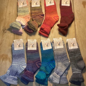 Könnte beinhalten: Eine Auswahl an bunten Wollsocken mit Etiketten, präsentiert auf einer Holzoberfläche. Die Socken weisen verschiedene Muster und Farbtöne auf, darunter Blau, Lila, Rot und Grün. Jede Socke hat einen umgeschlagenen Bund und ein Etikett mit einem Schafbild.
