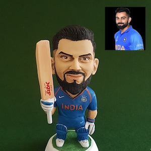 Pode incluir: Uma pequena figura de um jogador de cr&iacute;quete segurando um bast&atilde;o. A figura est&aacute; vestindo uma camisa azul com a palavra "INDIA" na frente. A figura est&aacute; em p&eacute; sobre uma base branca com a palavra "KOHLI" escrita nela.