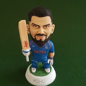 Pode incluir: Uma figura de um jogador de cr&iacute;quete segurando um bast&atilde;o. O jogador est&aacute; vestindo uma camisa azul com a palavra "INDIA" na frente. A base da figura &eacute; branca com a palavra "KOHLI" escrita nela.