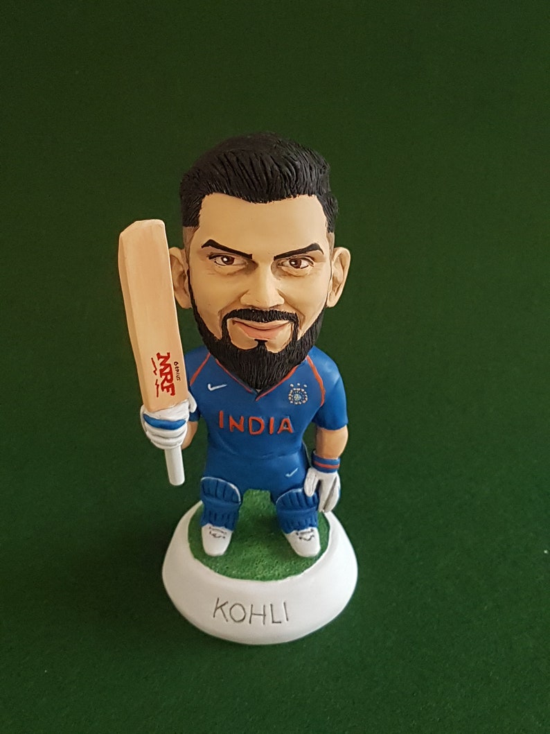 Pode incluir: Uma figura de um jogador de cr&iacute;quete segurando um bast&atilde;o. O jogador est&aacute; vestindo uma camisa azul com a palavra "INDIA" na frente. A base da figura &eacute; branca com a palavra "KOHLI" escrita nela.