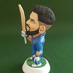 Pode incluir: Uma figura azul e branca de um jogador de cr&iacute;quete segurando um bast&atilde;o de madeira. A figura est&aacute; em p&eacute; sobre uma base branca com a palavra "HILI" escrita nela.