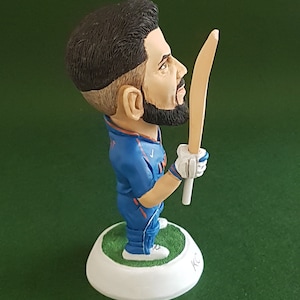 Pode incluir: Uma figura de um jogador de cr&iacute;quete azul e laranja segurando um taco de madeira. A figura est&aacute; em p&eacute; sobre uma base branca com grama verde ao redor da parte inferior. A base &eacute; assinada com "KC".