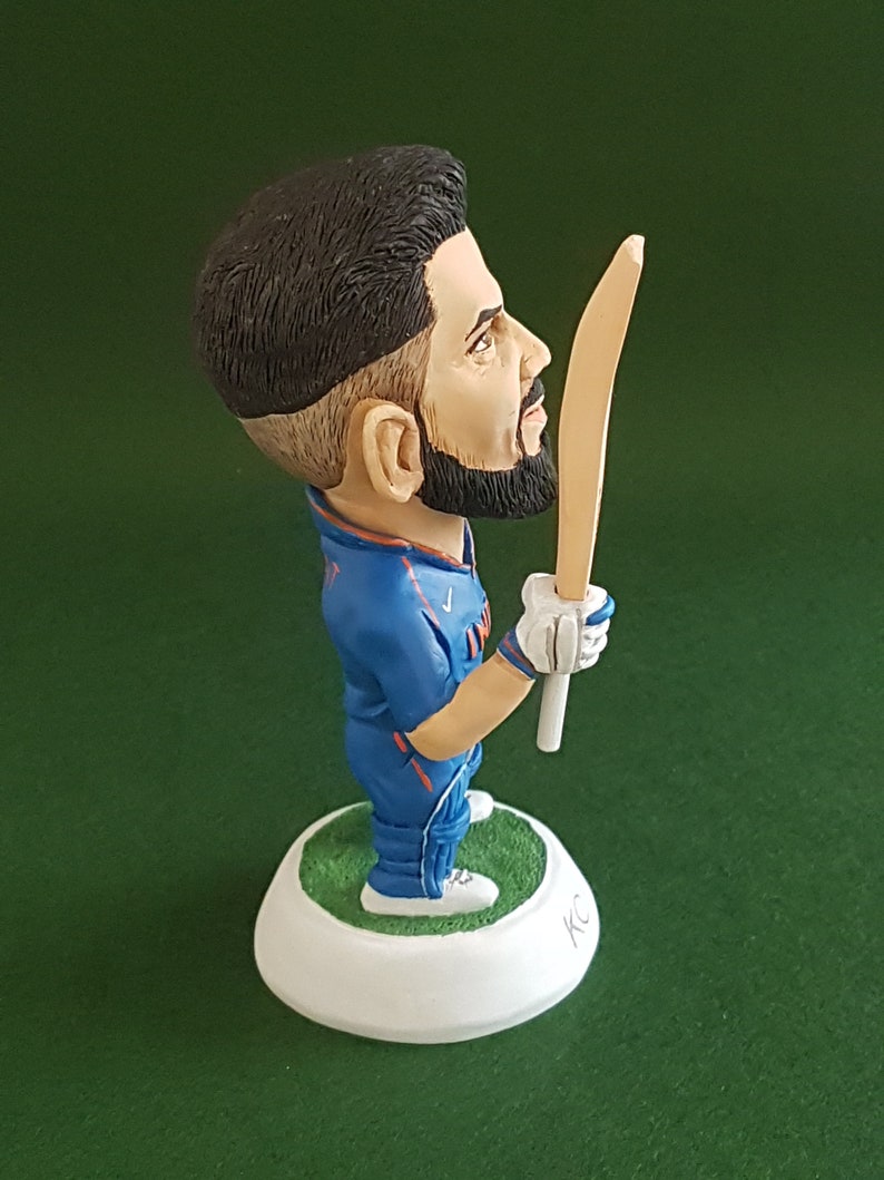 Pode incluir: Uma figura de um jogador de cr&iacute;quete azul e laranja segurando um taco de madeira. A figura est&aacute; em p&eacute; sobre uma base branca com grama verde ao redor da parte inferior. A base &eacute; assinada com "KC".