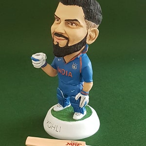 Pode incluir: Uma pequena figura de um jogador de cr&iacute;quete vestindo um uniforme azul e branco com a palavra "INDIA" no peito. A figura est&aacute; segurando uma pequena x&iacute;cara na m&atilde;o direita e est&aacute; em p&eacute; sobre uma base branca com a palavra "KOHLI" escrita nela. Um taco de cr&iacute;quete de madeira marrom com a palavra "MRF" impressa nele est&aacute; deitado sobre uma superf&iacute;cie verde.