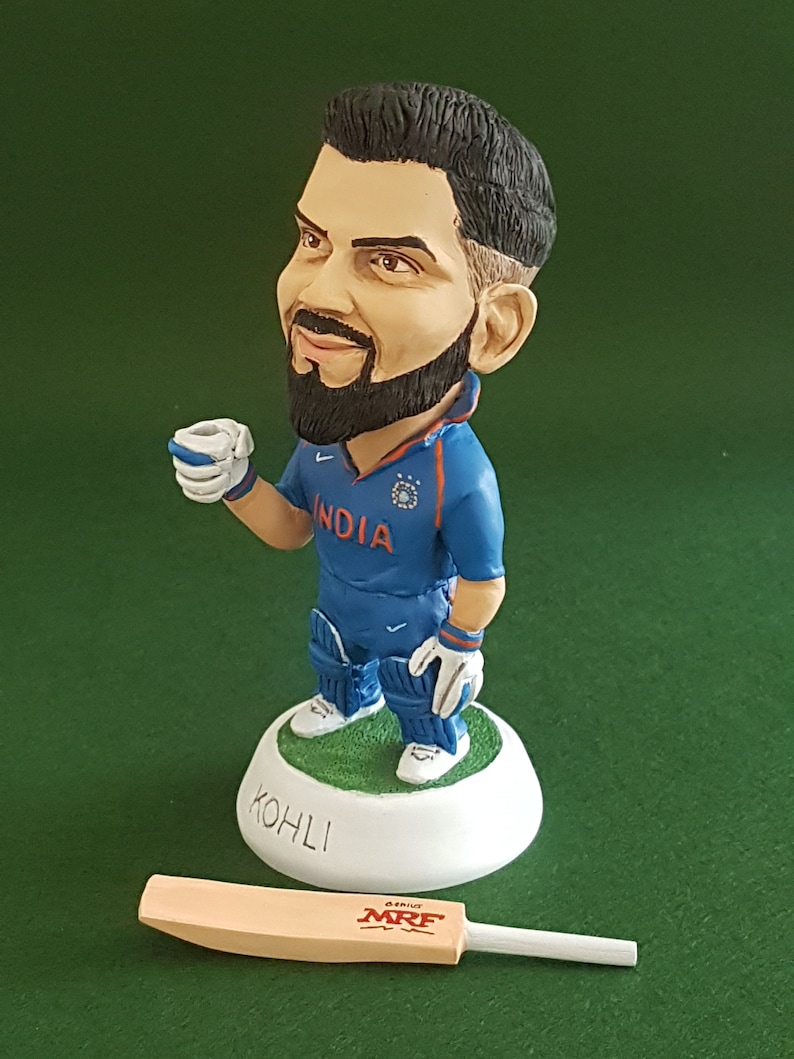 Pode incluir: Uma pequena figura de um jogador de cr&iacute;quete vestindo um uniforme azul e branco com a palavra "INDIA" no peito. A figura est&aacute; segurando uma pequena x&iacute;cara na m&atilde;o direita e est&aacute; em p&eacute; sobre uma base branca com a palavra "KOHLI" escrita nela. Um taco de cr&iacute;quete de madeira marrom com a palavra "MRF" impressa nele est&aacute; deitado sobre uma superf&iacute;cie verde.