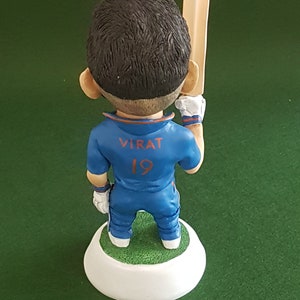 Pode incluir: Uma figura azul e laranja de um jogador de cr&iacute;quete segurando um bast&atilde;o de madeira. A figura est&aacute; vestindo uma camisa azul com o nome "VIRAT" e o n&uacute;mero "19" nas costas. A figura est&aacute; em p&eacute; sobre uma base branca.