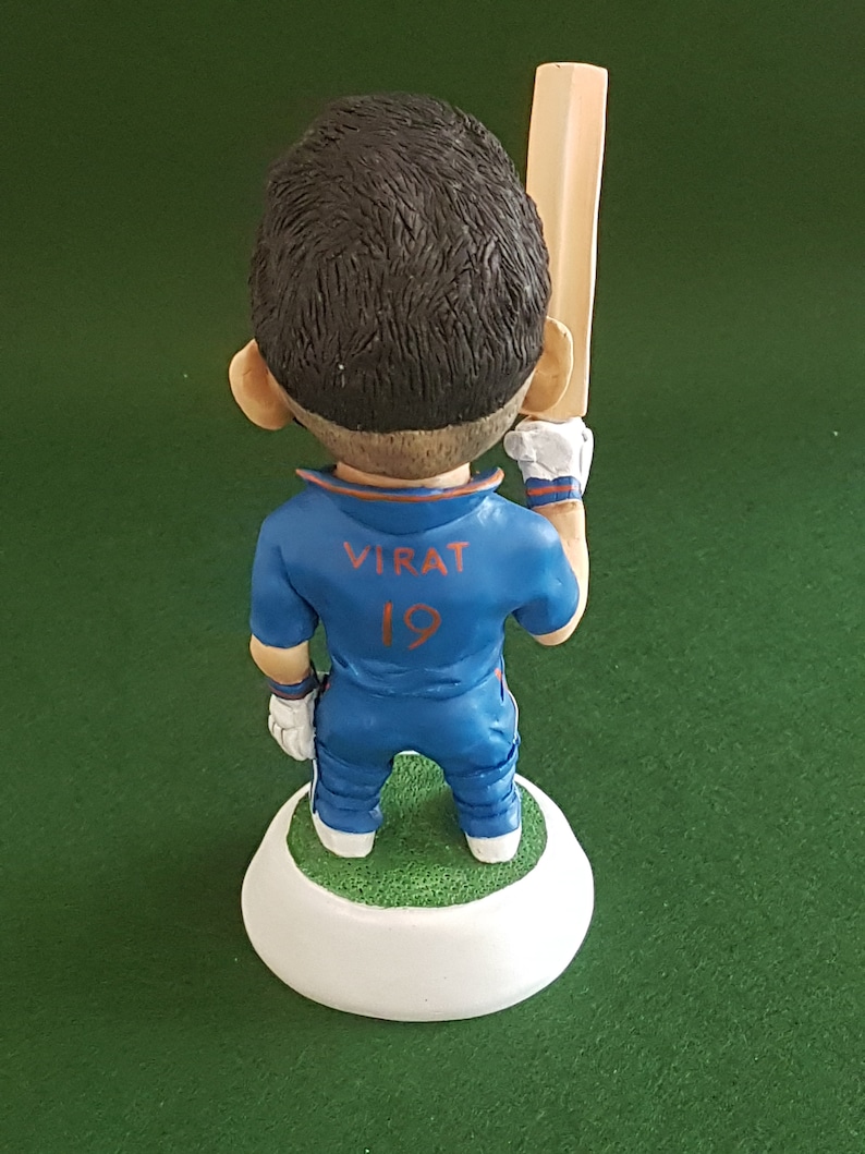 Pode incluir: Uma figura azul e laranja de um jogador de cr&iacute;quete segurando um bast&atilde;o de madeira. A figura est&aacute; vestindo uma camisa azul com o nome "VIRAT" e o n&uacute;mero "19" nas costas. A figura est&aacute; em p&eacute; sobre uma base branca.