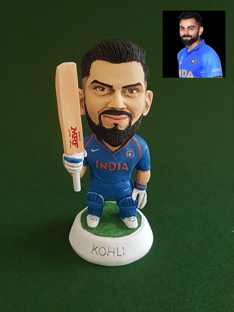 Pode incluir: Uma pequena figura de um jogador de cr&iacute;quete segurando um bast&atilde;o. A figura est&aacute; vestindo uma camisa azul com a palavra "INDIA" na frente. A figura est&aacute; em p&eacute; sobre uma base branca com a palavra "KOHLI" escrita nela.