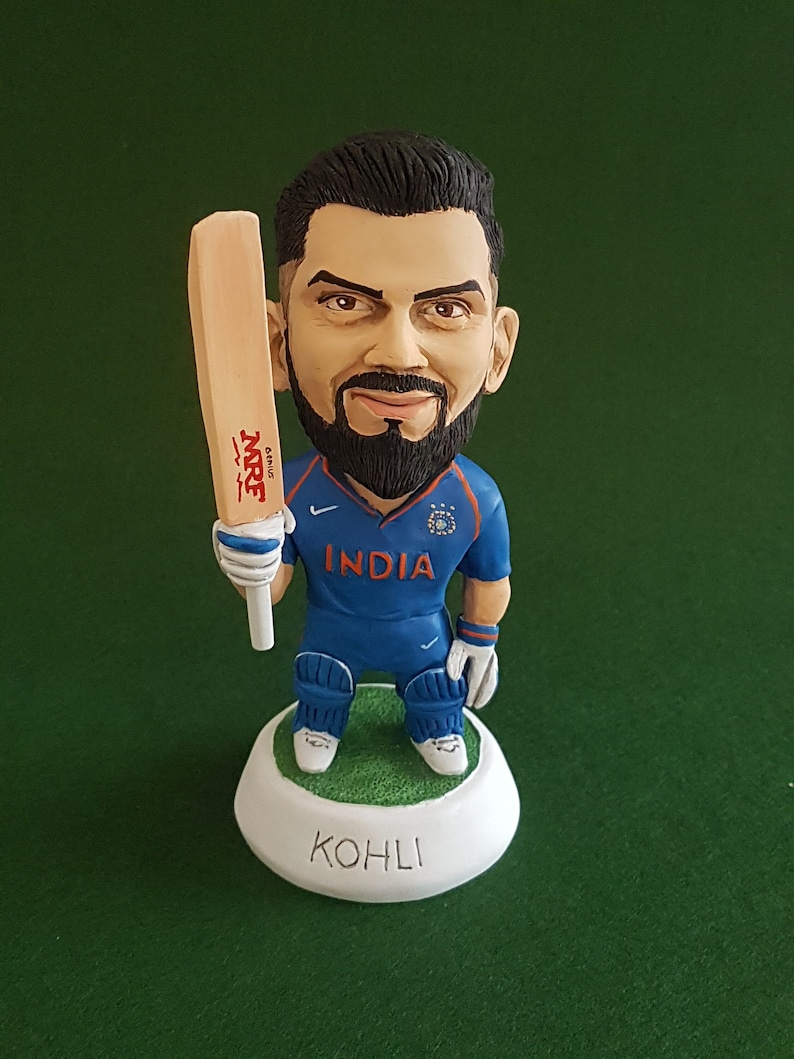 Pode incluir: Uma figura de um jogador de cr&iacute;quete segurando um bast&atilde;o. O jogador est&aacute; vestindo uma camisa azul com a palavra "INDIA" na frente. A base da figura &eacute; branca com o nome "KOHLI" escrito nela.