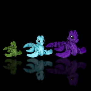 Peut inclure: Trois petites figurines de dragon articul&eacute;es en plastique de diff&eacute;rentes couleurs. Le dragon &agrave; gauche est vert, le dragon du milieu est bleu et le dragon &agrave; droite est violet.