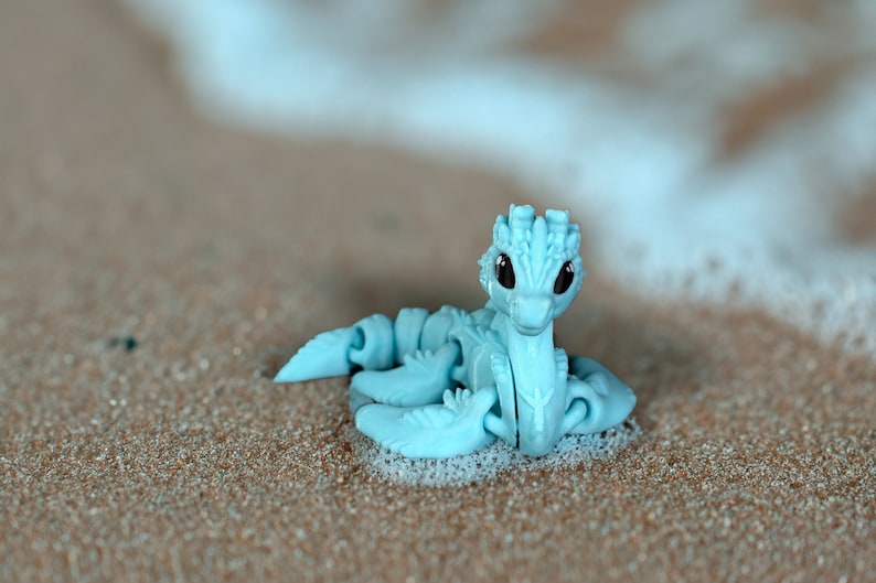 Peut inclure: Un dragon en plastique bleu clair avec des membres articul&eacute;s, assis sur une plage de sable avec de la mousse blanche.