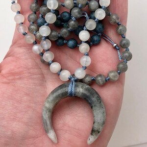 May include: A handcrafted necklace featuring a crescent moon pendant and a beaded strand. The necklace has a light blue cord and is adorned with white, gray, and blue beads. The moon pendant is gray with white accents.