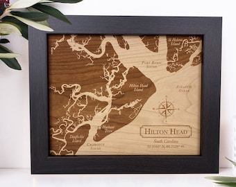 May include: A framed wooden map of Hilton Head, South Carolina, with a compass rose and the coordinates 32.2163° N, 80.7320° W. The map is engraved with the names of various bodies of water, including Calibogue Sound, Port Royal Sound, and the Atlantic Ocean.