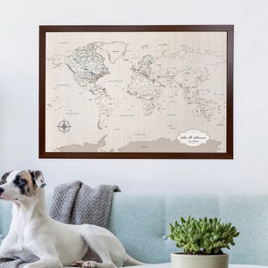 A cotton world push pin map with cotton texture on 100% cotton canvas. It is framed and hanging on a wall above a dog sitting on a sofa. The world map is neutral and natural tones and available personalized. It can be painted with watercolor paints.