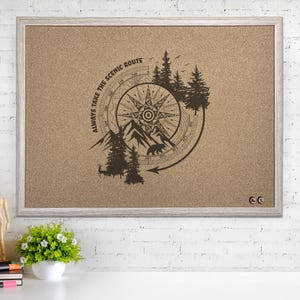 May include: A rectangular corkboard with a light wood frame. The board features a compass design with mountains, trees, and a bear, and the text "ALWAYS TAKE THE SCENIC ROUTE". The board is displayed on a white surface.