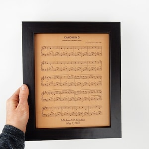 A person is holding up a framed leather sheet music engraving. The sheet music is a popular wedding song. The leather sheet music is also personalized to show a sample of how that looks with names and a date engraved at the bottom.