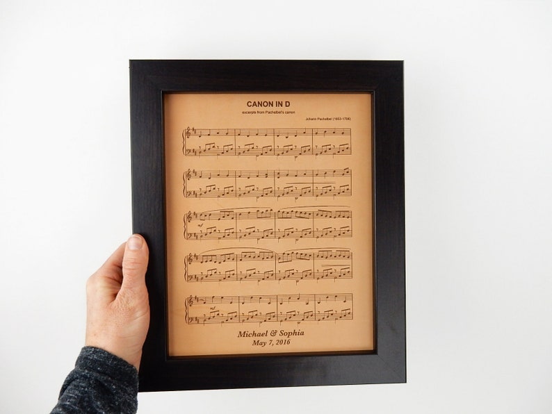 A person is holding up a framed leather sheet music engraving. The sheet music is a popular wedding song. The leather sheet music is also personalized to show a sample of how that looks with names and a date engraved at the bottom.