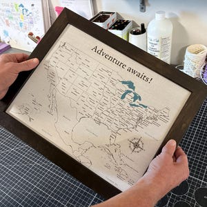 Finished 11x14 framed USA push pin travel map from JW Design Studio Gifts, held up to show its size and professional print quality. Each personalized cotton canvas travel map is designed, printed, cut, and assembled in-house by Jessica and Mike.