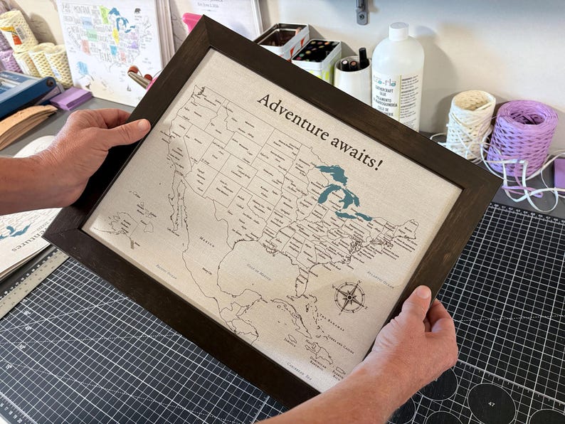Finished 11x14 framed USA push pin travel map from JW Design Studio Gifts, held up to show its size and professional print quality. Each personalized cotton canvas travel map is designed, printed, cut, and assembled in-house by Jessica and Mike.