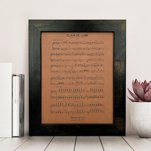 Peut inclure: Une impression encadrée de la partition de la chanson "Clair de Lune" de Claude Debussy. L'impression est sur du papier brun et a un cadre noir. Les noms "Mason & Ava" et la date "20 janvier 2023" sont imprimés en bas de la partition.