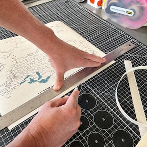 Behind the scenes at JW Design Studio Gifts: Jessica and Mike cut a custom 11x14 USA push pin travel map printed on cotton canvas, preparing each personalized map for framing and shipment in their home studio.