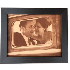 A leather photo of a wedding day celebration of a couple in a vintage car window. The leather photo is a warm sepia tone with rustic old fashioned look to it. The frame is a dark walnut frame.