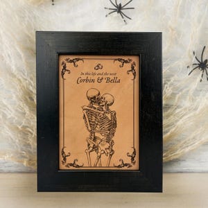 Op de afbeelding: Een ingelijst kunstwerk met een zwart houten frame. Het kunstwerk toont een omhelzend skeletpaar, met de tekst "In this life and the next Corbin & Bella" erboven. De achtergrond is lichtbruin.