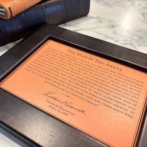 The Man in the Arena leather quote, framed. Styled sitting on a marble desktop with vintage book and leather journal nearby. The perfect gift for the distinguished gentlemen.