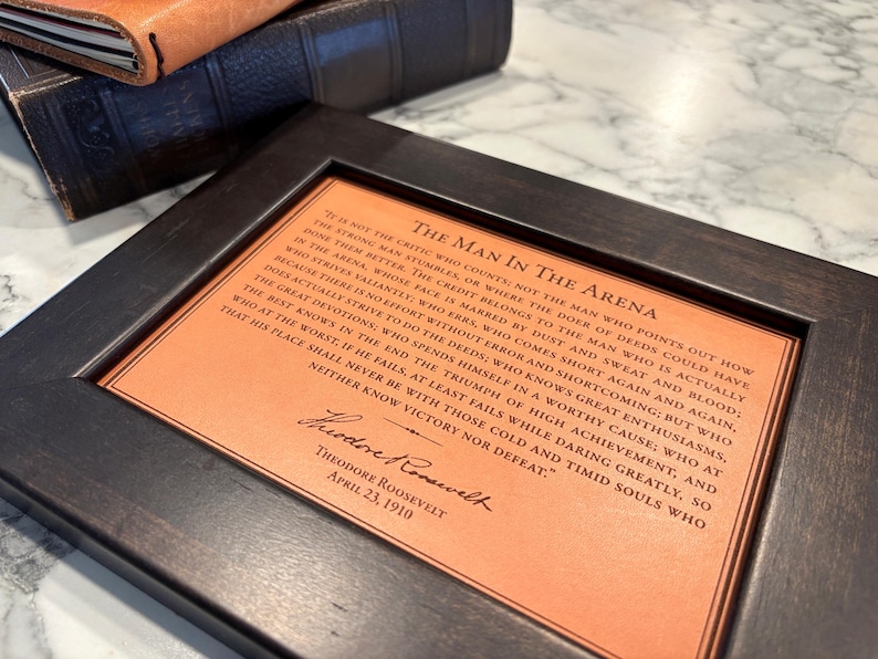 The Man in the Arena leather quote, framed. Styled sitting on a marble desktop with vintage book and leather journal nearby. The perfect gift for the distinguished gentlemen.