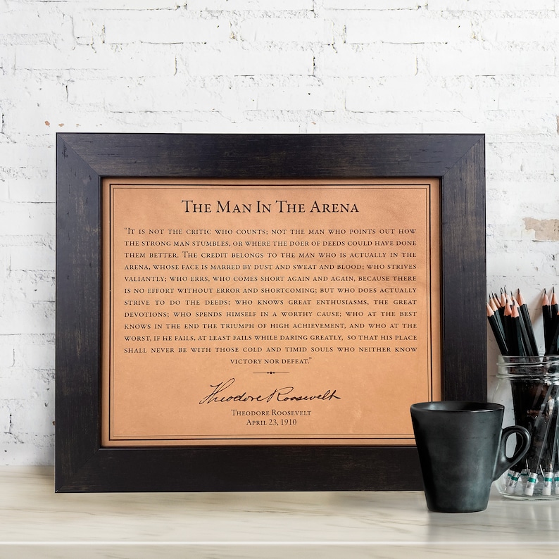 The Man in the Arena quote by Theodore Roosevelt is laser engraved on a leather for a luxury leather gift. It is framed in a dark walnut frame and sitting on a home office desk. This leather engraving is the perfect gift for graduation.