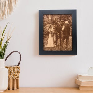 A engraved leather photo is framed and hanging on a wall in a boho styled room. The leather photo is of an outdoor wedding, very rustic and country themed wedding. It is framed in a dark walnut frame.