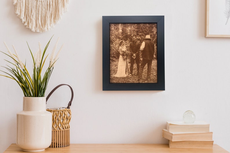 A engraved leather photo is framed and hanging on a wall in a boho styled room. The leather photo is of an outdoor wedding, very rustic and country themed wedding. It is framed in a dark walnut frame.