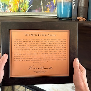 lifestyle image of a framed, the Man in the Arena quote engraved on leather. Man is holding the artwork in front of a wood mantle fireplace and vintage books.