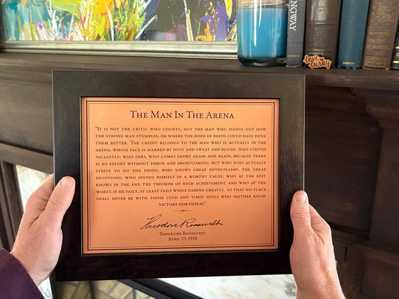 lifestyle image of a framed, the Man in the Arena quote engraved on leather. Man is holding the artwork in front of a wood mantle fireplace and vintage books.