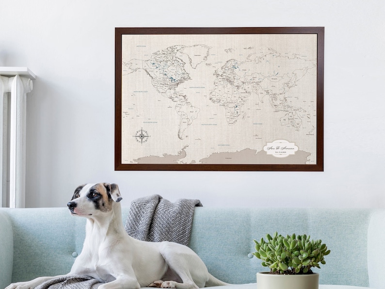 A cotton world push pin map with cotton texture on 100% cotton canvas. It is framed and hanging on a wall above a dog sitting on a sofa. The world map is neutral and natural tones and available personalized. It can be painted with watercolor paints.