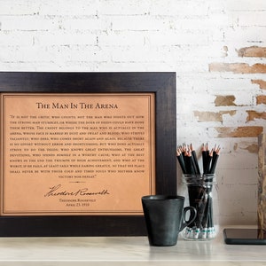 The iconic Man in the Arena quote by Teddy Roosevelt engraved on beautiful leather, presented as a high-end leather gift, showcased within a sophisticated dark walnut frame, adorning the desk of a professional lawyer