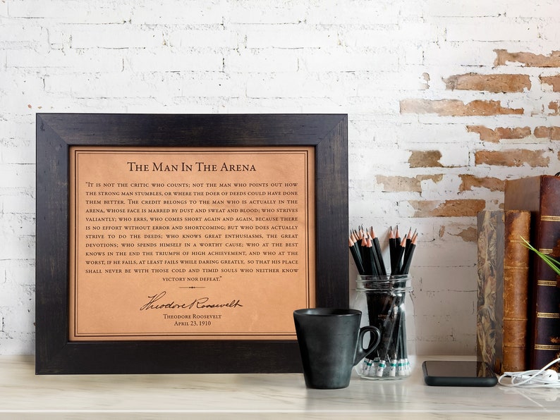 The iconic Man in the Arena quote by Teddy Roosevelt engraved on beautiful leather, presented as a high-end leather gift, showcased within a sophisticated dark walnut frame, adorning the desk of a professional lawyer