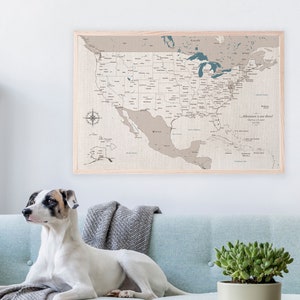 A USA cotton canvas travel map is framed and hanging on the wall. It has a neutral cream and beige tone with blue water details. A dog sits on a sofa in front of the map art in a modern home. The travel map is personalized with names and date.