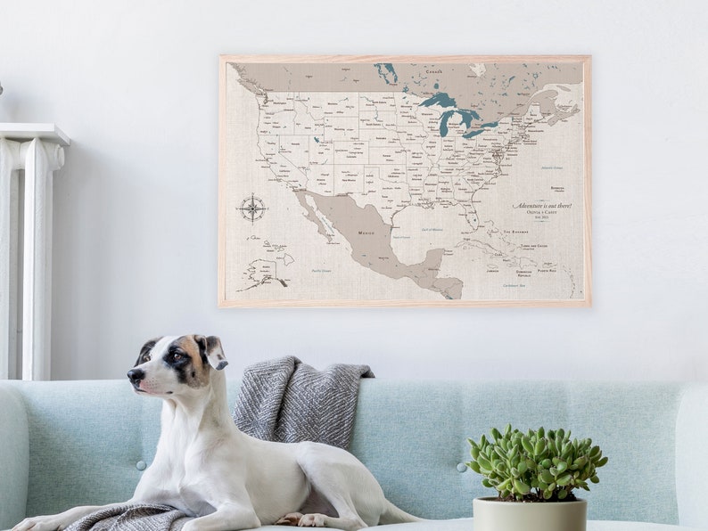 A USA cotton canvas travel map is framed and hanging on the wall. It has a neutral cream and beige tone with blue water details. A dog sits on a sofa in front of the map art in a modern home. The travel map is personalized with names and date.