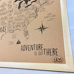 May include: A corkboard map of the United States with the text "Adventure is out there" printed below. The map is framed in wood and has a compass, a sailboat, and two push pins in the shape of a heart and an airplane.