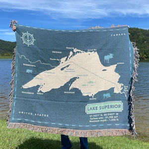May include: A woven tapestry throw blanket featuring a map of Lake Superior with details of the surrounding states and provinces. The map is in beige and white on a teal background. The text "LAKE SUPERIOR" is in white with the coordinates and measurements in black.