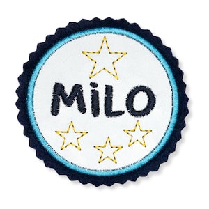 Könnte beinhalten: Ein weißer Filzkreis mit blauem Rand und gewellten Kanten. Der Kreis trägt den Namen "MILO" in schwarzer Stickerei und vier gelbe Sterne.